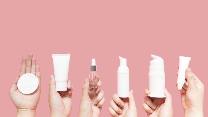 Skincare routine step for healthy skin - Woman hands holding facial cotton pad, foam, essential oil, serum, lotion and eye cream packaging on pink background. Beauty and cosmetic concept. Copy space.